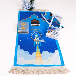 May include: A blue prayer mat with a rocket ship design, a small book, and a box with a ribbon. The mat has a fringe and the name "Amir" printed on it. The box has the words "Eid Mubarak" on it.