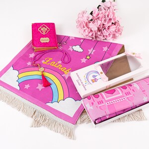 Puede incluir: Alfombra de oraci&oacute;n rosa con un dise&ntilde;o de arco&iacute;ris y estrellas, un libro de oraci&oacute;n rosa con el nombre "Zainab" en &eacute;l, una alfombra de oraci&oacute;n rosa en una caja y un collar de cuentas rosas con un dije dorado.