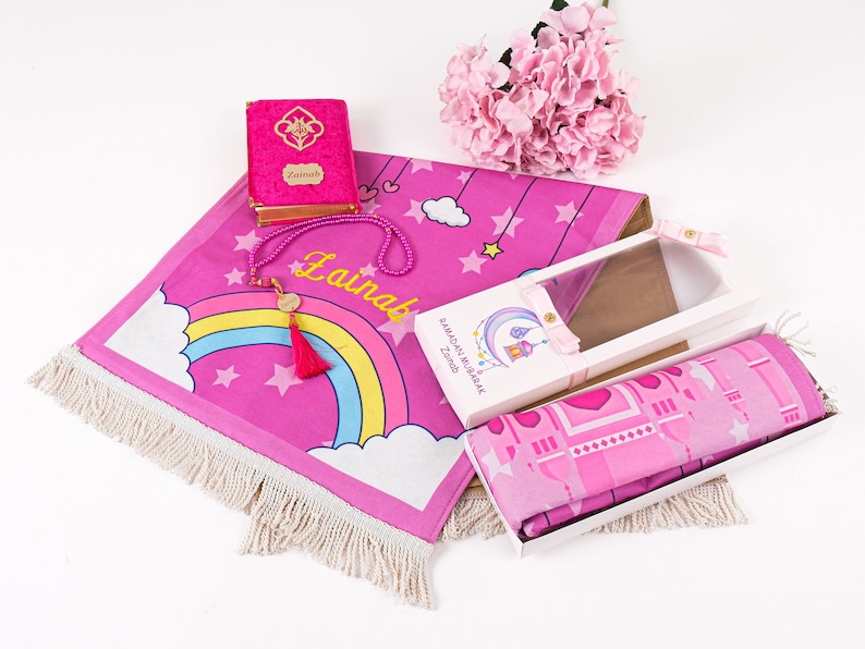 Puede incluir: Alfombra de oraci&oacute;n rosa con un dise&ntilde;o de arco&iacute;ris y estrellas, un libro de oraci&oacute;n rosa con el nombre "Zainab" en &eacute;l, una alfombra de oraci&oacute;n rosa en una caja y un collar de cuentas rosas con un dije dorado.