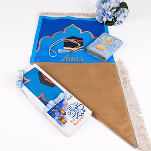 May include: A prayer mat set with a blue and tan color scheme. The set includes a prayer mat, a small Quran, and a beaded prayer necklace. The prayer mat has a design of the Kaaba and the name "Amir". A gift box with "Eid Mubarak" text is also included.
