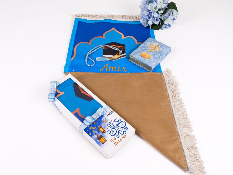 May include: A prayer mat set with a blue and tan color scheme. The set includes a prayer mat, a small Quran, and a beaded prayer necklace. The prayer mat has a design of the Kaaba and the name "Amir". A gift box with "Eid Mubarak" text is also included.