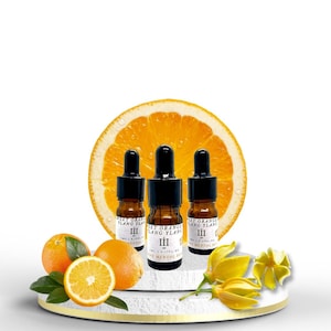 Op de afbeelding: Drie kleine amberkleurige glazen flesjes met zwarte druppelaars, gevuld met etherische olie. De flesjes zijn gelabeld met "Sweet Orange Ylang Ylang III" en zijn in een rij op een wit oppervlak gerangschikt. De flesjes zijn omringd door sinaasappelschijfjes en gele bloemen.