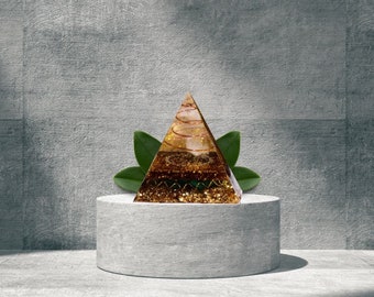May include: A gold and green crystal pyramid with a copper wire spiral inside, sitting on a gray concrete pedestal. The pyramid is surrounded by green leaves.