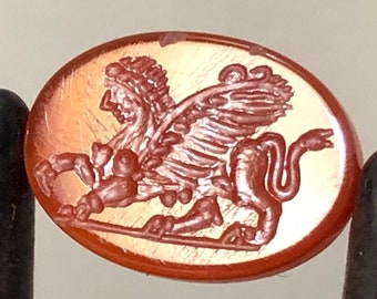 May include: A red carnelian gemstone cameo featuring a detailed carving of a winged lion. The cameo is oval shaped and has a smooth, polished surface.