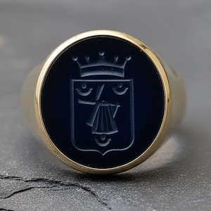 Op de afbeelding: Een gouden zegelring met een donkerblauwe, cirkelvormige face. De face heeft een gedetailleerd wapenontwerp. De ring is geplaatst tegen een donkere, getextureerde ondergrond, die het contrast tussen goud en donkerblauw benadrukt.