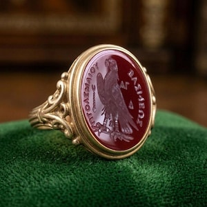 Pode incluir: Um anel de ouro com uma pedra vermelha ovalada com uma águia gravada e texto grego. O anel tem um design ornamentado em filigrana na banda e ao redor da configuração da pedra. O anel é exibido em uma superfície de veludo verde.