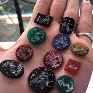 Puede incluir: Una colección de 11 cabujones de piedra preciosa tallados redondos y cuadrados con varios diseños. Los cabujones son en tonos de negro, verde, rojo, azul y marrón. Los diseños incluyen una medusa, un grifo, un lirio de flor, una abeja, un ancla y un escudo de armas.