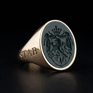 May include: A gold signet ring with an oval, dark green stone featuring a detailed crest with a crown, lion, and other heraldic elements. The word "STAB" is engraved on the side of the ring.