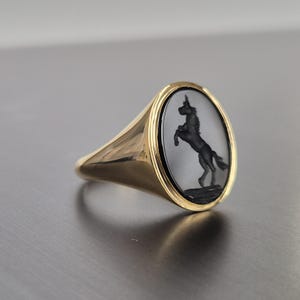 Op de afbeelding: Gouden zegelring met een ovale voorkant. De ring heeft een zwart eenhoornontwerp tegen een grijze achtergrond. De ringband is glad en gepolijst.