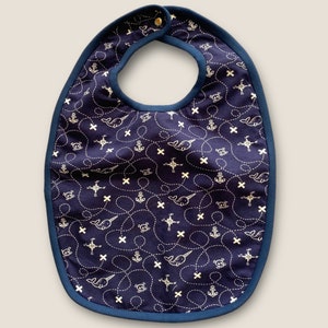 Puede incluir: Babero azul marino para bebé con un estampado náutico. El babero presenta un diseño repetido de ballenas blancas, anclas, brújulas y líneas punteadas sobre un fondo azul oscuro. Tiene un ribete azul oscuro y un cierre de botón a presión en el cuello.