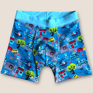 Puede incluir: Calzoncillos bóxer azules con un estampado lúdico de una escena urbana. El diseño incluye casas, coches, helicópteros, árboles y policías. La cintura es de color azul claro liso.