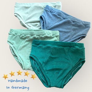 May include: A set of four pairs of underwear in varying shades of blue and green, including light mint, sage, denim blue, and dark forest green. The garments feature a textured fabric, elastic waistbands, and five yellow stars with the text Handmade In Germany.