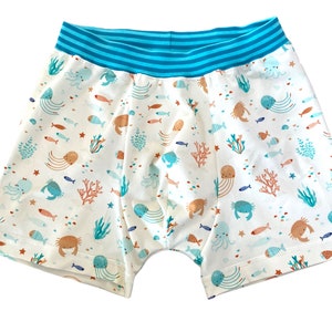 Op de afbeelding: Boxershorts met een witte basis en een herhalend patroon van kleurrijke zeedieren, waaronder krabben, kwallen en vissen, samen met koraal en zeewier. Het kledingstuk heeft een brede, geribbelde tailleband met horizontale strepen in felblauw.