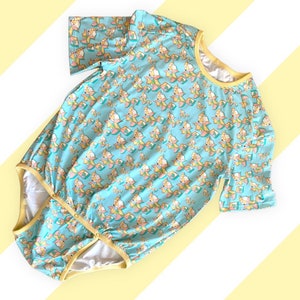 May include: A light blue onesie with a repeating geometric fox pattern in yellow, pink, and teal. The onesie has short sleeves, a yellow trim, and snap closures at the crotch.