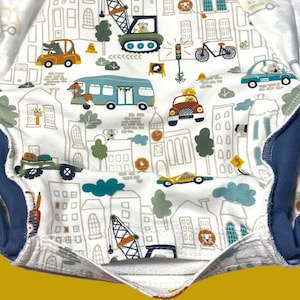 May include: White fabric with a blue trim, featuring a repeating pattern of a city scene with buildings, cars, and trees. The pattern is in shades of gray, brown, and green.