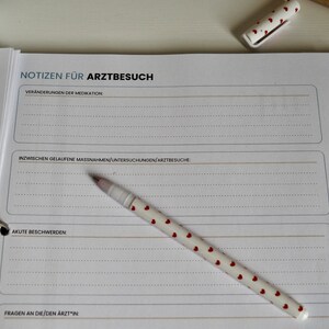 May include: A white notebook with a black binder ring and a pen with red hearts on it. The notebook has the title "Notizen für Arztbesuch" and sections for "Veränderungen der Medikationen", "Zwischen gelaufene Massnahmen/Untersuchungen/Arztbesuche", "Akute Beschwerden", and "Fragen an die/den Arzt/in".