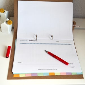 May include: A white binder with the text "SYMPTOM TRACKER" on the cover. The binder is open to a page with a grid for tracking symptoms. A red pen is lying on the page.