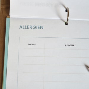 May include: A white page with the word "ALLERGIES" at the top, followed by the words "DATE" and "TRIGGER" in a table format. The page is from a notebook with a silver ring binder.