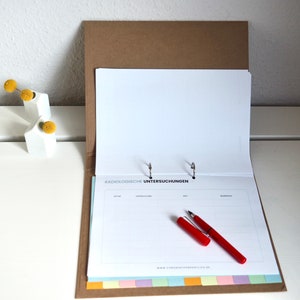 May include: A brown binder with a white page open, showing a gridded form with the text "RADIOLOGISCHE UNTERSUCHUNGEN" and a red pen lying on top. The binder is on a white surface.