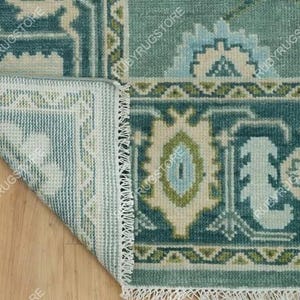 May include: Close-up of the edge of a teal blue rug with a white fringe. The rug has a geometric pattern with green, white, and blue accents. The rug is folded over to show the back side, which is white with a green and white pattern.