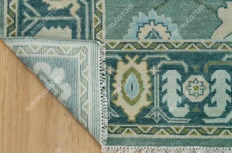 May include: Close-up of the edge of a teal blue rug with a white fringe. The rug has a geometric pattern with green, white, and blue accents. The rug is folded over to show the back side, which is white with a green and white pattern.