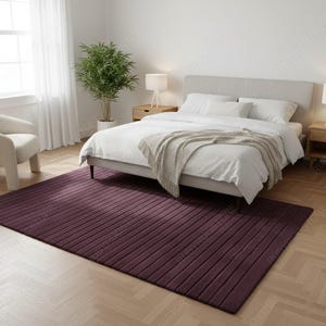 May include: A deep purple rectangular rug with a ribbed texture is placed on a wooden floor in a bedroom setting. The rug is positioned in front of a bed with white bedding and a light gray headboard. A white chair and a potted plant are also visible.