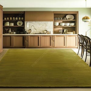 Puede incluir: Una gran alfombra rectangular verde oliva domina el primer plano de una cocina. La textura de la alfombra es sutilmente visible. La cocina cuenta con gabinetes de madera, una placa para salpicaduras de mármol y una mesa de comedor con sillas. El estilo es moderno y minimalista.
