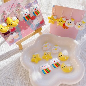May include: A selection of quirky earrings and charms, including yellow moons, stars, and white clouds with rainbow details. The earrings are presented on pink cards, and the charms are in a white, cloud-shaped dish. The earrings have silver hooks.
