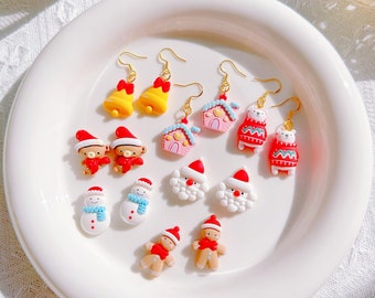 May include: A selection of festive Christmas earrings on a white plate. Designs include bells, houses, bears, snowmen, Santa faces, and gingerbread men. The earrings are in red, white, yellow, and pink, with gold-coloured hooks.