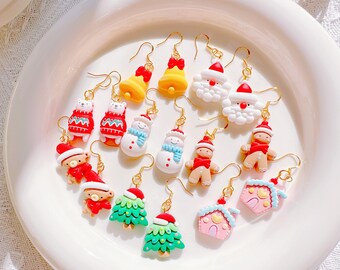 May include: A selection of festive Christmas earrings featuring Santa Claus, snowmen, gingerbread people, Christmas trees, and houses. The earrings are brightly coloured resin with gold-tone hooks. They are presented on a white plate.