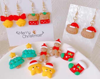 May include: A collection of Christmas-themed earrings and charms. The earrings feature designs like bells with red bows, teddy bears in Santa hats, and Christmas trees. Charms include gingerbread men, stars, and stockings. The text "Merry Christmas" is visible.