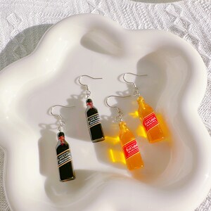 May include: Earrings featuring miniature bottle designs. Two pairs of earrings are visible, one with black bottles labeled "Black Label" and the other with orange bottles labeled "Red Label". The earrings are displayed on a white, wavy-edged plate.