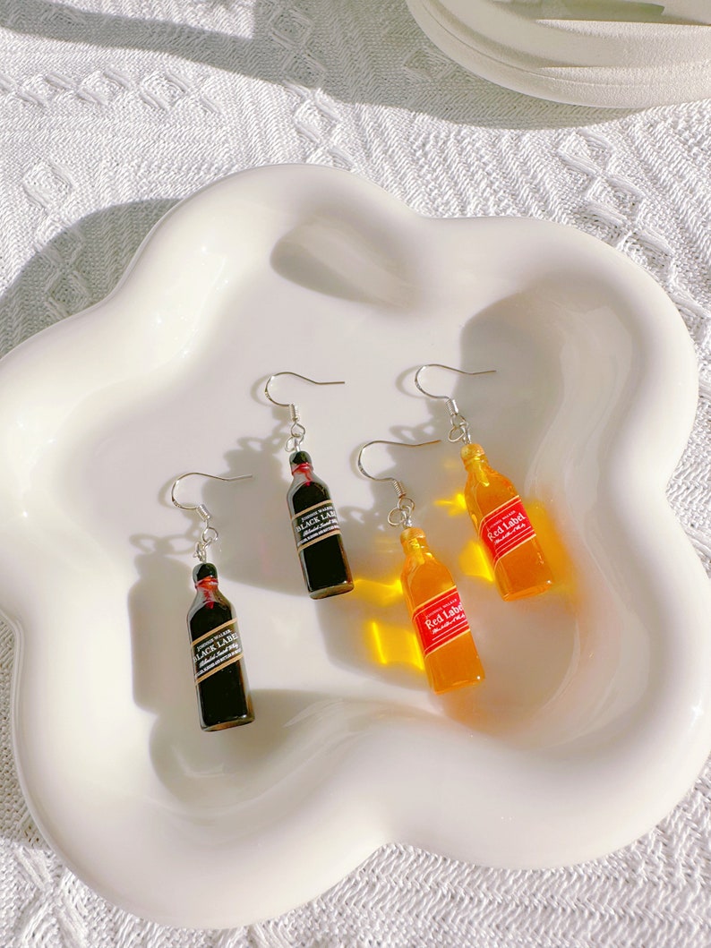 May include: Earrings featuring miniature bottle designs. Two pairs of earrings are visible, one with black bottles labeled "Black Label" and the other with orange bottles labeled "Red Label". The earrings are displayed on a white, wavy-edged plate.