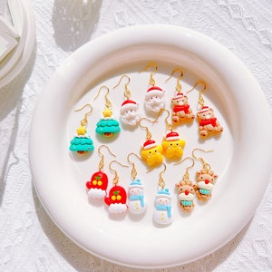 May include: A collection of Christmas-themed earrings displayed on a white plate. The earrings feature various festive designs, including Santa Claus, Christmas trees, snowmen, reindeer, and teddy bears, all with gold-colored hooks.