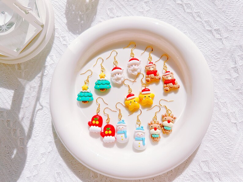 May include: A collection of Christmas-themed earrings displayed on a white plate. The earrings feature various festive designs, including Santa Claus, Christmas trees, snowmen, reindeer, and teddy bears, all with gold-colored hooks.