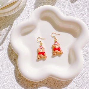 May include: A pair of festive bear earrings. Each earring features a small, tan bear wearing a red scarf and a Santa hat. The earrings have gold-colored hooks and are displayed on a white, flower-shaped dish.