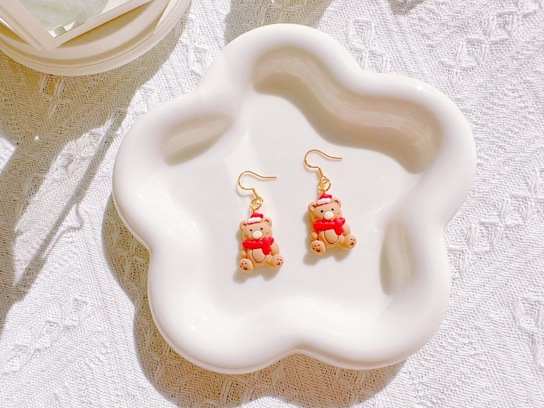 May include: A pair of festive bear earrings. Each earring features a small, tan bear wearing a red scarf and a Santa hat. The earrings have gold-colored hooks and are displayed on a white, flower-shaped dish.