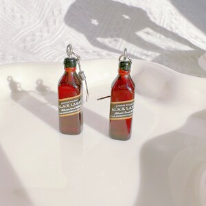 May include: Miniature brown bottle earrings with a black and gold label that reads "Johnnie Walker Black Label". The earrings have a silver-colored metal loop and a small metal rod. The bottles have a green cap.