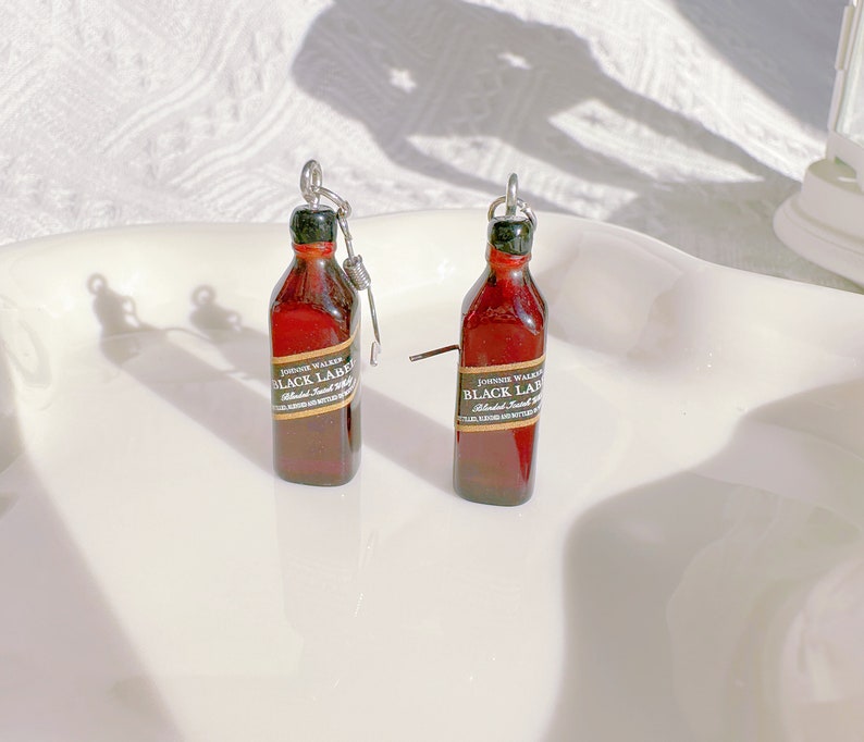 May include: Miniature brown bottle earrings with a black and gold label that reads "Johnnie Walker Black Label". The earrings have a silver-colored metal loop and a small metal rod. The bottles have a green cap.