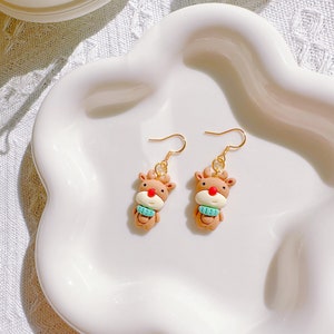 May include: A pair of reindeer-shaped earrings. Each earring features a brown reindeer with a red nose and a light blue scarf. The earrings have gold-colored hooks and are displayed on a white, scalloped plate.