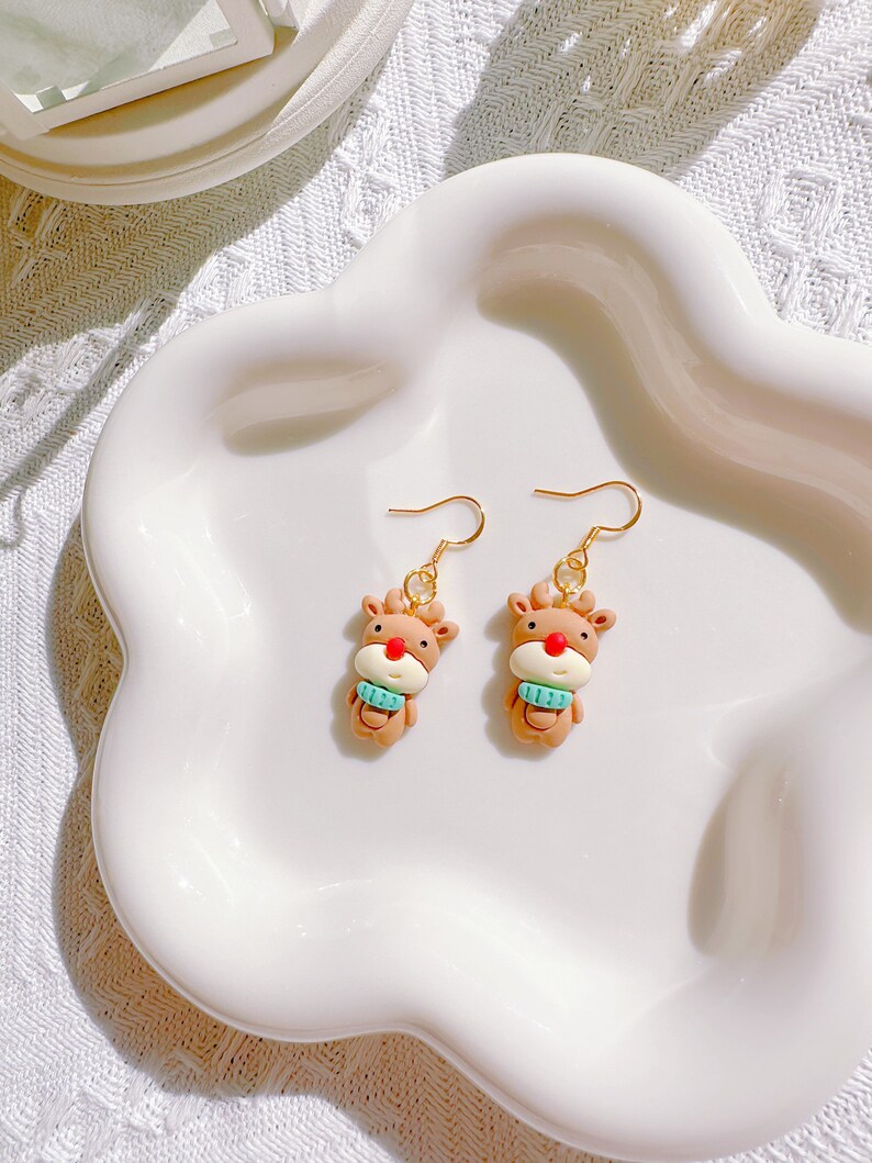 May include: A pair of reindeer-shaped earrings. Each earring features a brown reindeer with a red nose and a light blue scarf. The earrings have gold-colored hooks and are displayed on a white, scalloped plate.