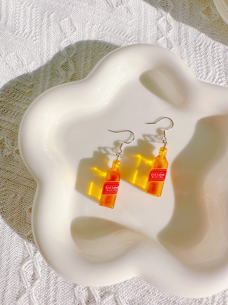 May include: A pair of novelty earrings shaped like miniature whiskey bottles with the text "Red Label" on the label. The earrings are orange and yellow, with silver-colored ear wires. They are displayed on a white, heart-shaped dish.
