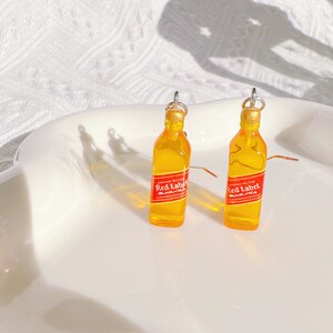 May include: A pair of novelty earrings shaped like miniature whiskey bottles. The earrings are yellow with red and white labels that read "Red Label". The bottle tops are gold, and the earrings have silver hooks.