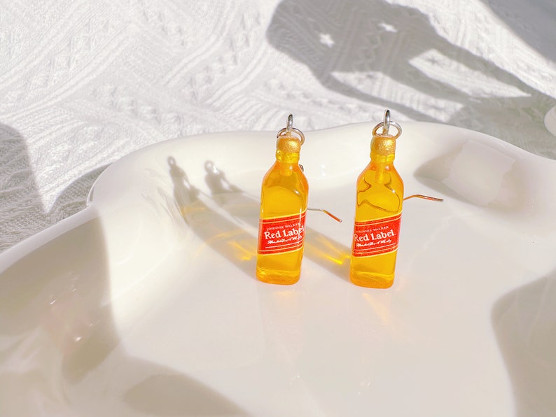 May include: A pair of novelty earrings shaped like miniature whiskey bottles. The earrings are yellow with red and white labels that read "Red Label". The bottle tops are gold, and the earrings have silver hooks.