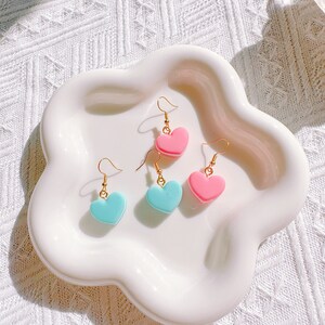 Pode incluir: Quatro brincos em forma de coração, rosa e azul claro, com ganchos dourados, exibidos em um prato branco com bordas onduladas. Os brincos são dispostos em pares, mostrando seu design simétrico e cores vibrantes.