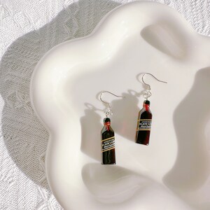 May include: A pair of novelty earrings featuring miniature replicas of Johnnie Walker Black Label whiskey bottles. The earrings have silver-colored fish hook ear wires and are set against a white background.