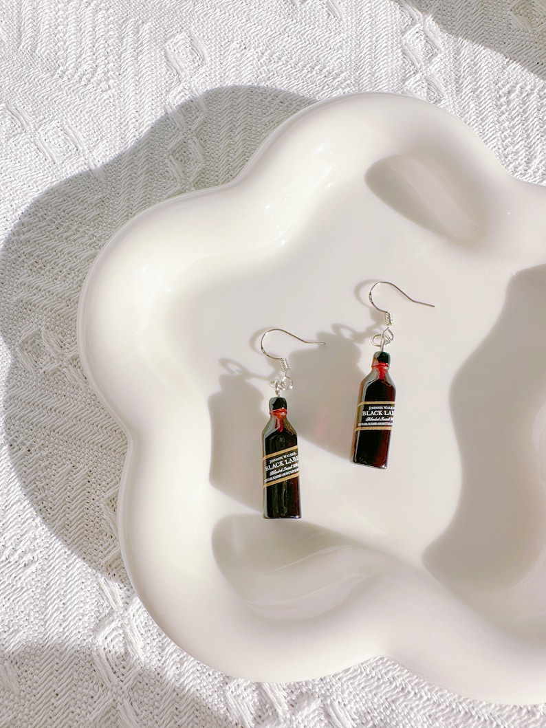 May include: A pair of novelty earrings featuring miniature replicas of Johnnie Walker Black Label whiskey bottles. The earrings have silver-colored fish hook ear wires and are set against a white background.