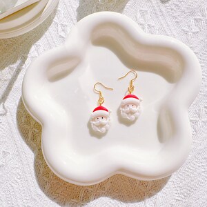 May include: A pair of Santa Claus earrings with gold-colored hooks. Each earring features a miniature Santa head with a red hat, white beard, and rosy cheeks. The earrings are displayed on a white, cloud-shaped dish.