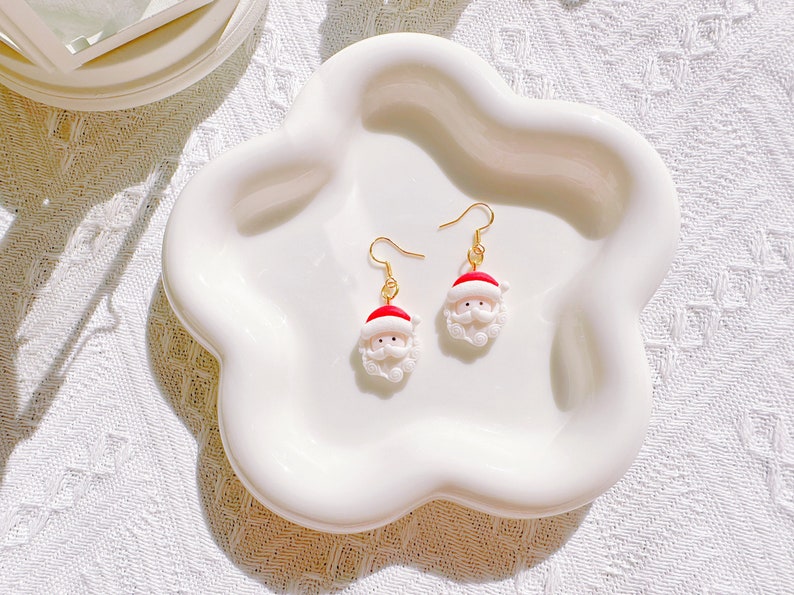 May include: A pair of Santa Claus earrings with gold-colored hooks. Each earring features a miniature Santa head with a red hat, white beard, and rosy cheeks. The earrings are displayed on a white, cloud-shaped dish.