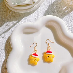 May include: A pair of gold-colored hook earrings. Each earring features a yellow star-shaped charm wearing a red and white Santa hat. The earrings are displayed on a white, wavy-edged plate.